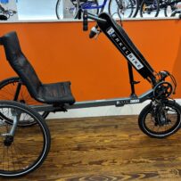 Side view of a used Berkel Bike Pro adaptive hand‑and‑foot cycle showing the crankset, pedals, and front wheel against a wood floor.
