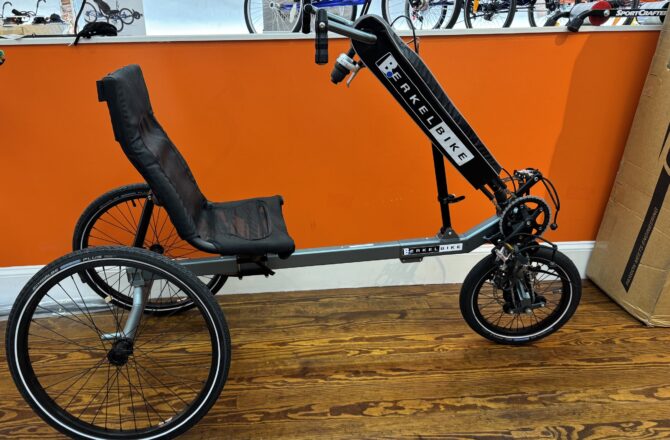 Side view of a used Berkel Bike Pro adaptive hand‑and‑foot cycle showing the crankset, pedals, and front wheel against a wood floor.